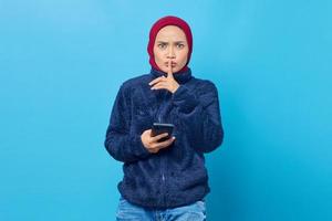 Hermosa joven asiática sosteniendo un teléfono móvil y hace un gesto de silencio sobre fondo azul. foto