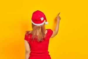 Young woman wearing Christmas hat in back position pointing finger at copy space on yellow background photo