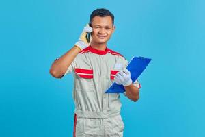 Sonriente joven mecánico asiático hablando por móvil y sosteniendo el portapapeles aislado sobre fondo azul. foto