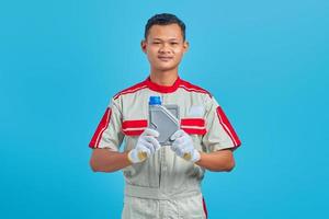 Retrato de sonriente joven mecánico asiático mostrando una botella de plástico de aceite de motor en la mano aislado sobre fondo azul.