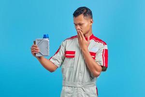 Retrato de joven mecánico asiático conmocionado sosteniendo una botella de plástico de aceite de motor y cubriendo la boca sobre fondo azul manos