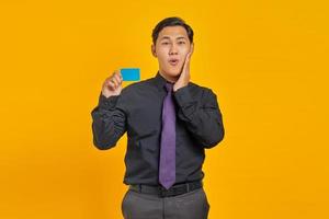 Happy young Asian businessman holding credit card and touching cheek on yellow background photo