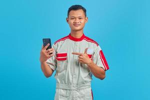 Portrait of smiling young mechanic pointing finger at mobile phone isolated on blue background photo