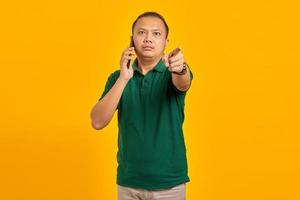 Angry Asian young man talking on smartphone and doing warning sign isolated over yellow background photo