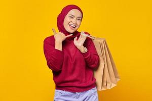 Beautiful Asian woman showing phone call gesture and holding shopping bags on yellow background photo