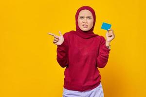 Conmocionada mujer asiática sosteniendo una tarjeta de crédito mientras apunta con el dedo en el espacio de la copia sobre fondo amarillo foto