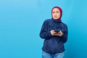 Confused young Asian woman using smartphone and looking sideways isolated on blue background