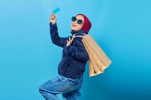 Emocionada y alegre joven asiática sosteniendo bolsas de compras y tarjeta de crédito sobre fondo azul. foto