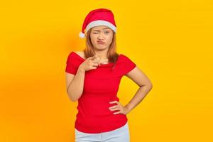 Portrait of sad young Asian woman with hands on waist and wearing Christmas hat on yellow background photo