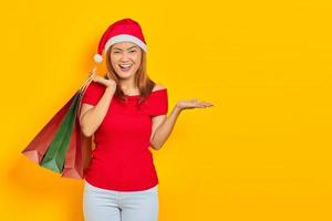 Cheerful young Asian woman in Santa Claus hat holding shopping bags and showing copy space in palms on yellow background