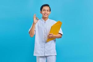 Portrait of cheerful asian male nurse holding yellow folder and making ok gesture on blue background photo
