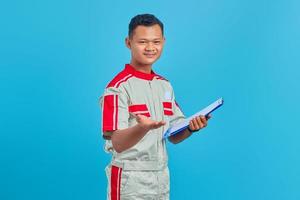 Portrait of a cheerful young mechanic holding clipboard and pointing with palm at camera isolated on blue background photo