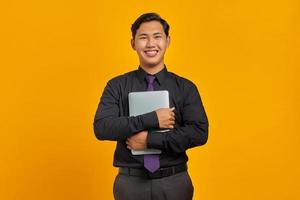 Smiling handsome young businessman hugging laptop and looking at camera on yellow background photo