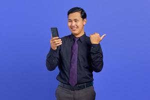 Cheerful handsome young businessman holding smartphone and pointing to copy space on purple background
