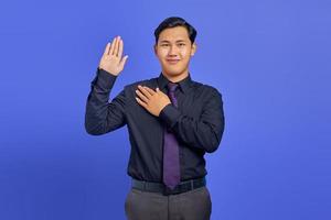 Smiling handsome young businessman with confident face swearing with hands on chest and open palms on purple background