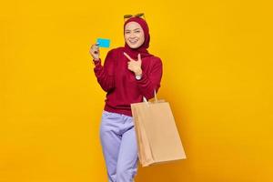 Portrait of cheerful Asian woman holding shopping bag and showing credit card over yellow background photo