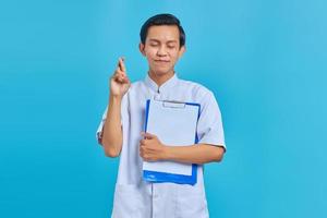 Portrait of happy young Asian male nurse crossing fingers and showing clipboard on blue background photo