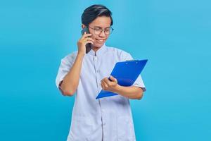 Portrait of male nurse smiling and talking on smartphone while holding clipboard isolated on blue background photo