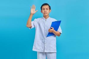 Portrait of cheerful male nurse holding clipboard standing and waving forward on blue background photo