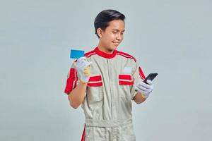 Portrait of Cheerful young mechanic showing credit card and using smartphone on gray background photo