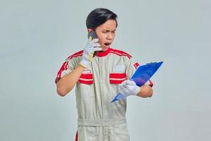 Portrait of young Asian mechanic talking on phone and holding clipboard with surprised expression isolated on gray background photo