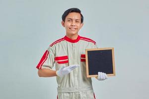 Portrait of cheerful young mechanic wearing uniform pointing to blank board with palms on gray background photo