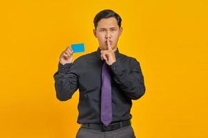 Portrait of handsome Asian businessman showing silent gesture and holding credit card on yellow background photo