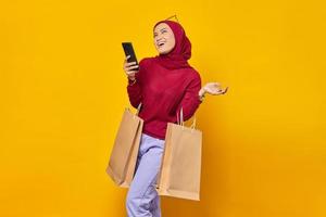 Smiling young Asian woman using a mobile phone and holding shopping bags on yellow background photo