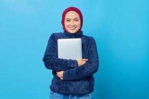 Photo of cheerful asian woman wearing blue jacket hugging laptop on chest and looking at camera