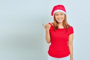 Photo of beautiful Asian girl in christmas hat with smiling facial expression and pointing aside on white background