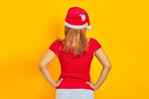 Young woman wearing Christmas hat in back position holding hands on waist confidently on yellow background