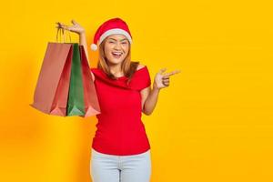 Cheerful young Asian woman in Santa Claus hat showing shopping bags and pointing finger at copy space on yellow background photo