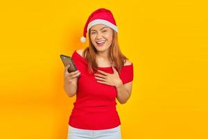 Excited young Asian woman in Santa Claus hat holding cell phone with hands on chest on yellow background