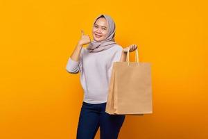 Retrato de mujer asiática sonriente sosteniendo la bolsa de compras y mostrando los pulgares hacia arriba sobre fondo amarillo foto