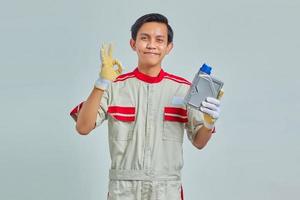 Retrato de smilling guapo vestido con uniforme de mecánico sosteniendo una botella de plástico de aceite de motor y mostrando aprobación con el pulgar hacia arriba sobre fondo gris