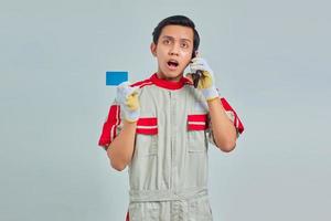 Shocked handsome mechanic talking on smartphone and holding credit card on gray background photo