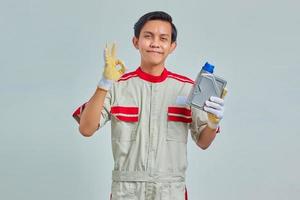 Retrato de smilling guapo vestido con uniforme de mecánico sosteniendo una botella de plástico de aceite de motor y mostrando aprobación con el pulgar hacia arriba sobre fondo gris