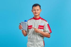Retrato de sonriente joven mecánico asiático apuntando a una botella de plástico de aceite de motor con el dedo sobre fondo azul.
