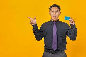 Conmocionado empresario asiático sosteniendo una tarjeta de crédito mientras apunta con el dedo en el espacio de la copia sobre fondo amarillo foto