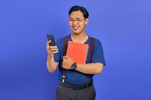 Handsome young student smiling looking at mobile phone and holding notebook on purple background