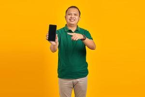 Sonriente joven guapo apuntando al teléfono inteligente con pantalla en blanco sobre fondo amarillo foto