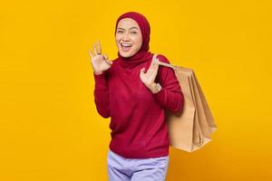 Portrait of smiling Asian woman holding shopping bags and making okay sign with fingers over yellow background photo