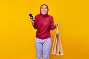 cheerful young Asian woman holding mobile phone and shopping bags on yellow background photo