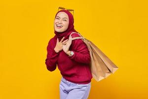 Happy young Asian woman with hand on a chest and holding shopping bags on yellow background