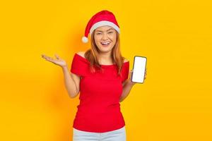 Smiling young Asian woman in Santa Claus hat extending hand inviting to come and showing smartphone on yellow background photo