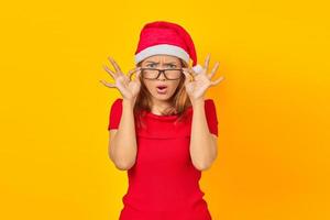 Surprised young Asian woman wearing Christmas hat with glasses while open mouth on yellow background