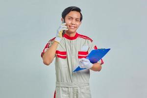 Retrato de alegre joven mecánico asiático hablando por teléfono y sosteniendo el portapapeles aislado sobre fondo gris foto