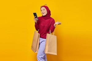 Smiling young Asian woman using a mobile phone and holding shopping bags on yellow background photo