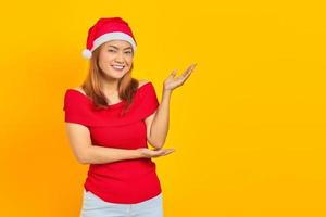 Retrato de hermosa niña asiática con sombrero de navidad apuntando al espacio vacío con la palma aislada sobre fondo amarillo foto