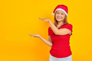 Smiling young Asian woman in christmas hat extending hand to the side inviting to come photo